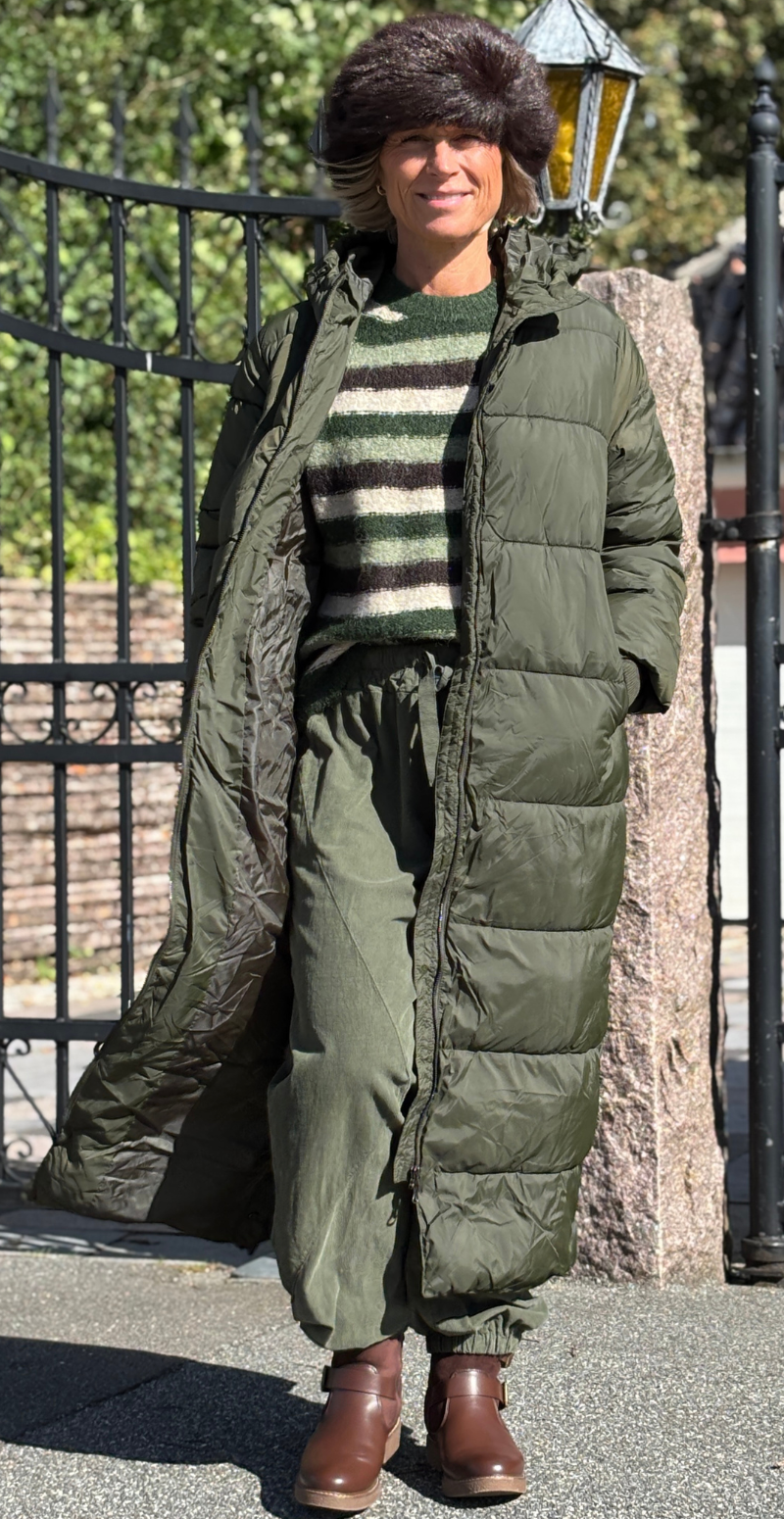 Kvinde set forfra i armygrøn Liva frakke med hætte, stribet sweater og matchende bukser.