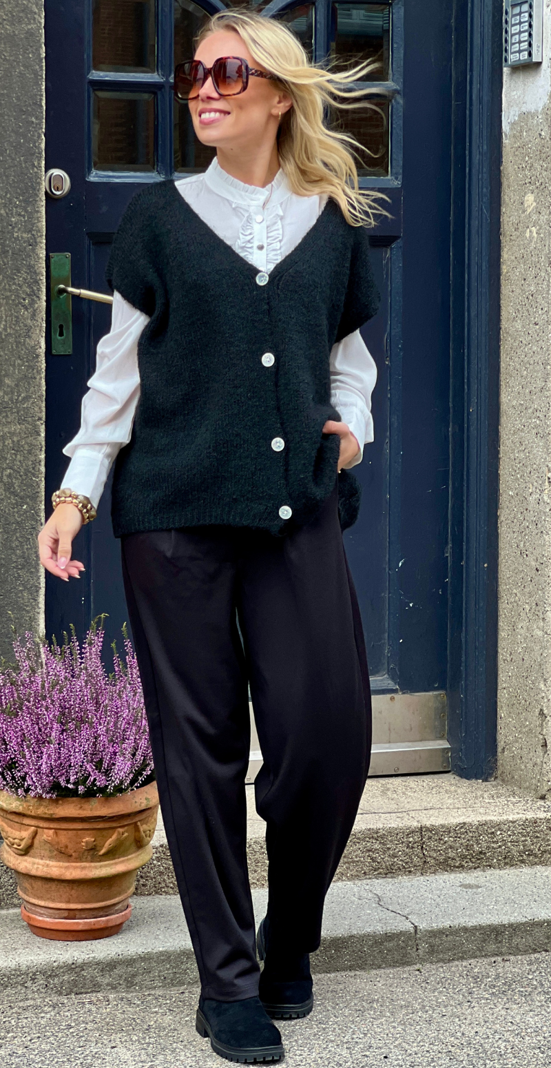 Kvinde i sorte Heika bukser, hvid bluse og sort strikket vest set forfra.
