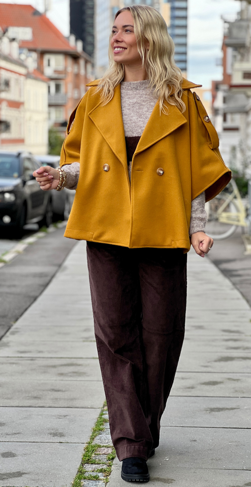 Kvinde i Cleo kort poncho jakke med knapper fortil i farven mustard fra LikeLondon.com set forfra.